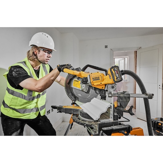 Worker using DEWALT DCS781N cordless mitre saw to cut trim in a room, wearing safety gear.