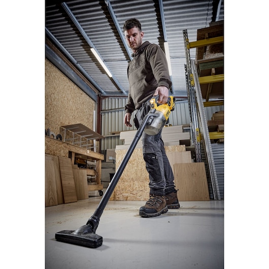 Person using a DEWALT DCV501LN vacuum cleaner on a workshop floor amid wood panels and shelves.