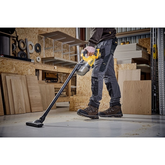 Person using DEWALT cordless vacuum to clean sawdust on workshop floor, surrounded by wood panels.