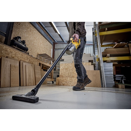 Person using a DEWALT cordless vacuum to clean a workshop floor with wooden boards and shelves visible.