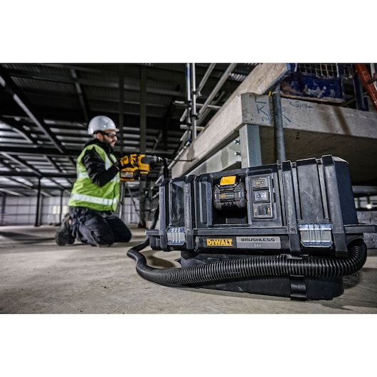 DEWALT DCV586MT2 brushless vacuum on a construction site floor, worker kneeling nearby in safety gear.