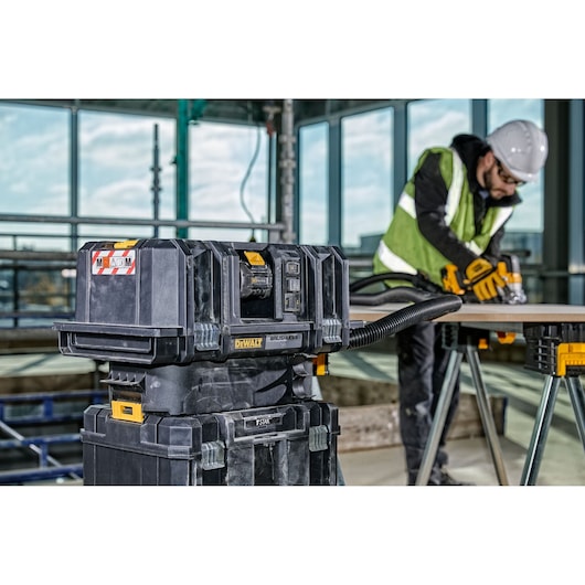 DEWALT DCV586MT2 dust extractor on site, worker in PPE sanding wood nearby, industrial setting.