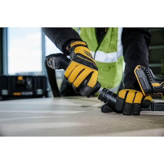 Hands with DEWALT gloves connect a vacuum hose to a power tool on a work surface, with a DEWALT case in background.