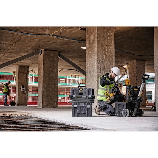 Worker in safety vest uses DEWALT equipment on a construction site with toolboxes and concrete pillars.