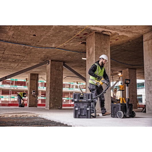 Worker uses DEWALT equipment at a construction site, near boxes and lighting, wearing a helmet and safety vest.