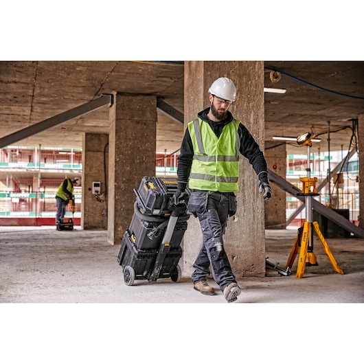 Worker with DEWALT rolling toolbox and light on tripod at construction site, wearing safety gear.