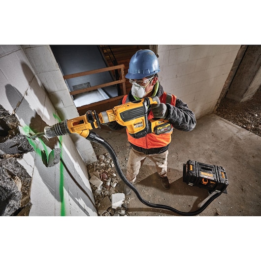 Worker using a DEWALT cordless vacuum and drill on a construction site wall with protective gear.