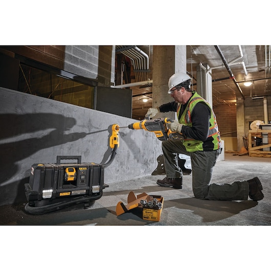 Worker uses DEWALT DCV586MT2 dust extractor and drill on concrete wall at construction site.