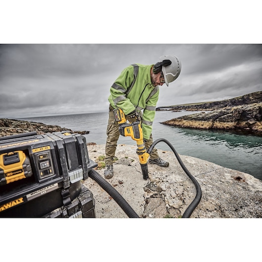 Worker uses yellow DEWALT tool outdoors, drilling concrete near water. Tool case and hose in foreground.