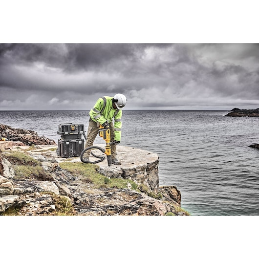 Worker using DEWALT tool on rocky cliff by the sea, with storage boxes nearby under cloudy sky.