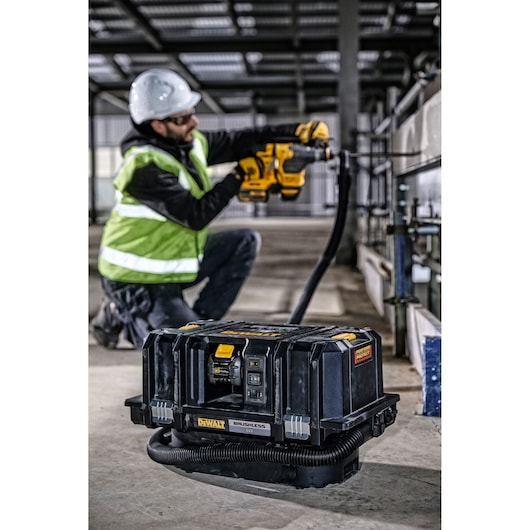 DEWALT DCV586MT2 vacuum on site; worker in safety gear uses a drill. Industrial setting with concrete floor.