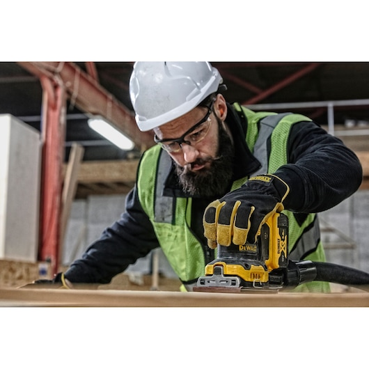 Worker uses a DEWALT DCW200P2 sander on wood, wearing gloves, helmet, and safety vest at a construction site.