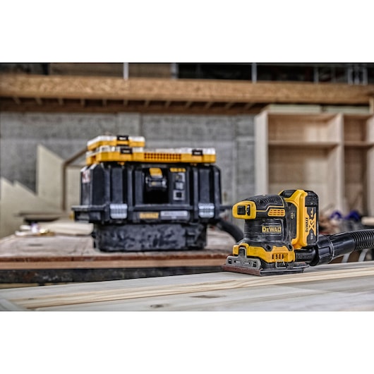 DEWALT cordless sander on wood, with storage box in background in a workshop setting.