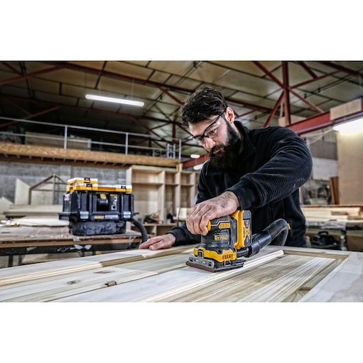 Person sanding wood with DEWALT sander DCW200P2 in workshop. Toolbox in background. Indoor setting.
