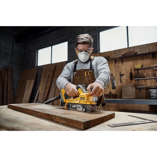A person in a workshop uses a DEWALT DCW220N yellow belt sander on a wooden plank. The person’s face is blurred. The surroundings show wooden panels and various tools.