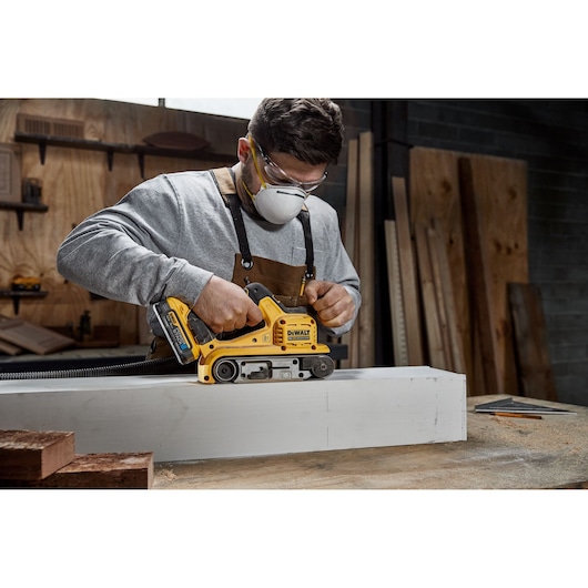 A person wearing an apron is using a yellow DEWALT DCW220N cordless belt sander to sand a large piece of wood in a workshop.