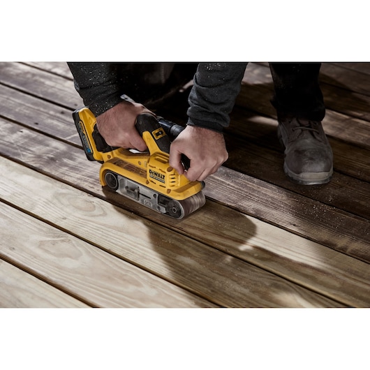 A person using a yellow DEWALT cordless belt sander on wooden boards to smooth their surface.