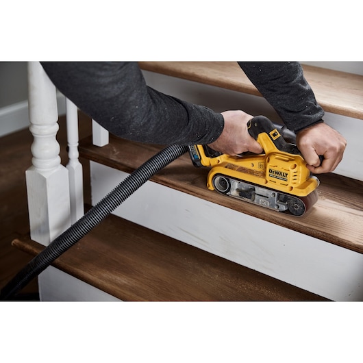 A person using a DEWALT DCW220N cordless belt sander to sand wooden stairs, with a vacuum hose attached to the tool for dust collection.