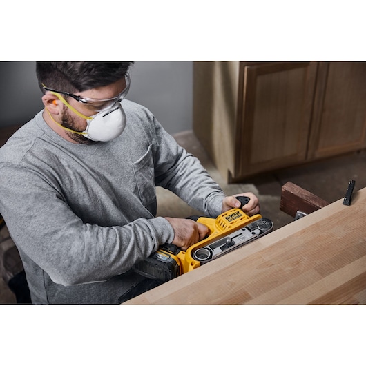 A person wearing safety gear uses a DEWALT DCW220N cordless belt sander to sand a wooden surface in a workshop.