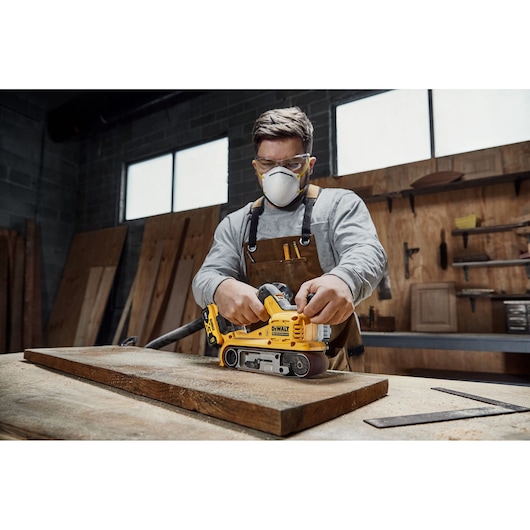 A person in a workshop using a DEWALT DCW220P2 cordless belt sander to smooth a wooden plank on a table. The person is wearing a brown apron and safety equipment, and the workspace has wooden boards and tools visible in the background.