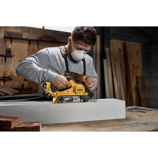 A person using a DEWALT cordless belt sander (SKU: DCW220P2) on a piece of wood in a workshop. The person is wearing safety goggles and an apron, and their face is blurred.