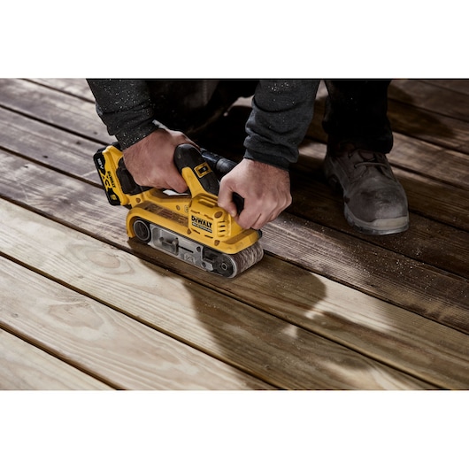 A person using a yellow DEWALT cordless belt sander (model DCW220P2) to sand wooden floorboards.