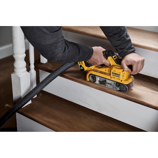 A person is using a DEWALT DCW220P2 yellow cordless belt sander to sand a wooden staircase step. A vacuum hose is attached to the tool for dust extraction.