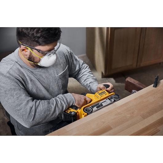 A person using a DEWALT DCW220P2 cordless belt sander to smooth the surface of a wooden table. The user is wearing safety goggles and a dust mask.