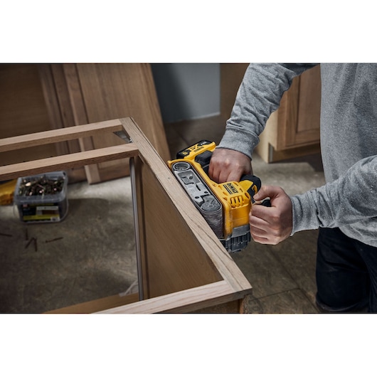 A person using a yellow DEWALT cordless belt sander to smooth the edge of a wooden frame in an indoor workspace.