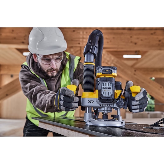Worker using DEWALT XR DCW620H2 cordless router on wood in construction site, wearing safety gear.