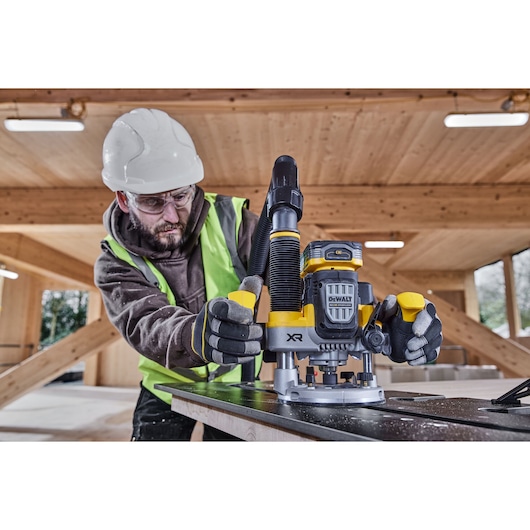 Worker uses DEWALT cordless router DCW620H2 on wood in a construction site, wearing safety gear and gloves.