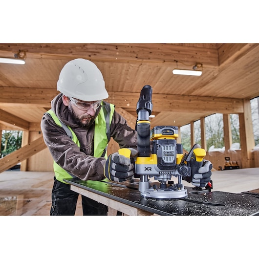 Worker uses a DEWALT DCW620H2 router on wood in a construction setting, wearing safety gear and gloves.
