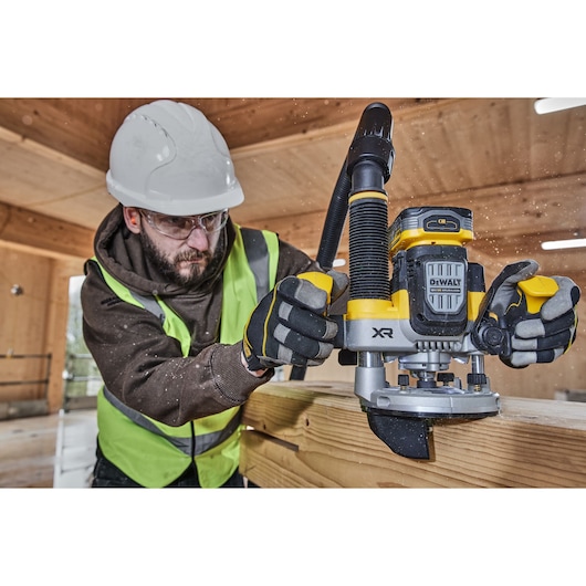 Worker uses DEWALT XR router on wood, wearing gloves, vest, and helmet in a workshop.