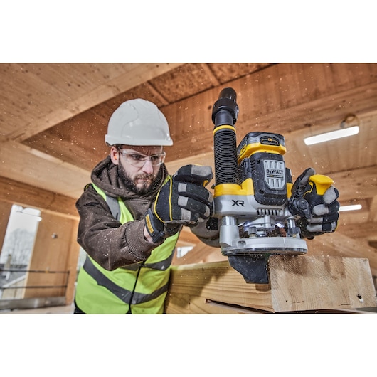 Worker uses DEWALT router DCW620H2 on wood, wearing safety gear in an indoor construction setting.