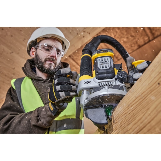 Person using DEWALT DCW620H2 router on wood. Worker wears safety gear while operating tool indoors.