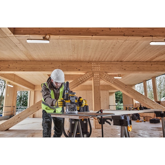 Worker uses DEWALT router on wood table inside a timber building, wearing helmet, gloves, and safety vest.