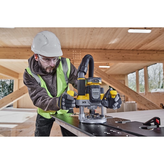 Worker uses DEWALT XR router DCW620NT on a metal surface in a wooden workshop, wearing safety gear.
