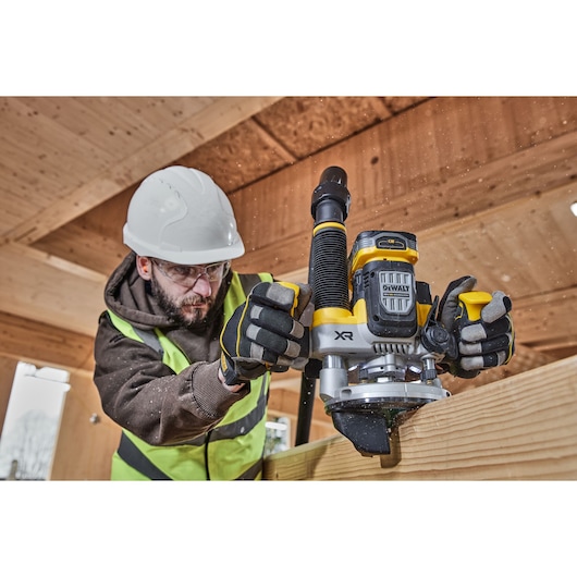 Worker uses a DEWALT DCW620NT router on wood; wearing gloves, vest, and helmet in a construction setting.