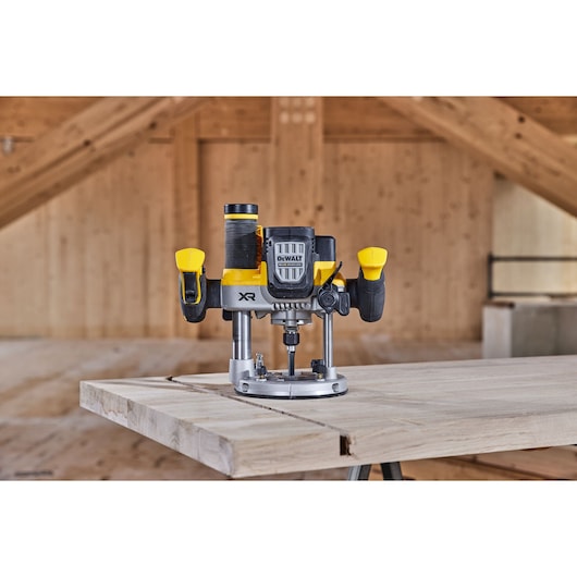 A DEWALT cordless plunge router positioned on a wooden workbench in a workshop setting. The tool features yellow and black handles and is designed for precision woodworking tasks.