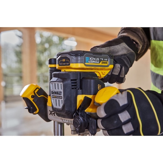 Gloved hands operate a DEWALT cordless router with Powerstack battery in a workshop setting.