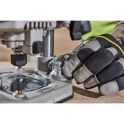 Gloved hand adjusts bolts on a DEWALT router tool base placed on a wooden surface.