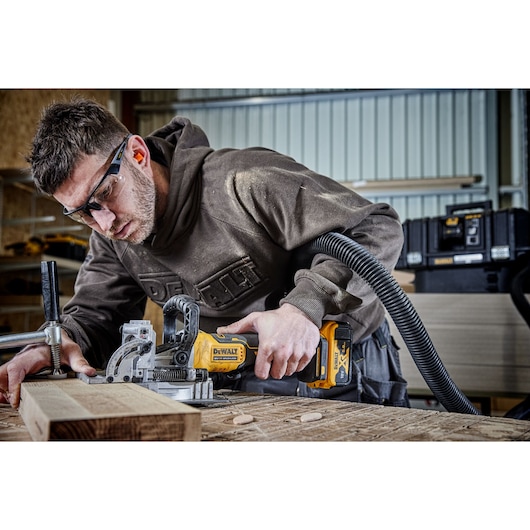 Person uses DEWALT cordless joiner to cut wood in a workshop, with dust extractor attached and toolboxes in background.
