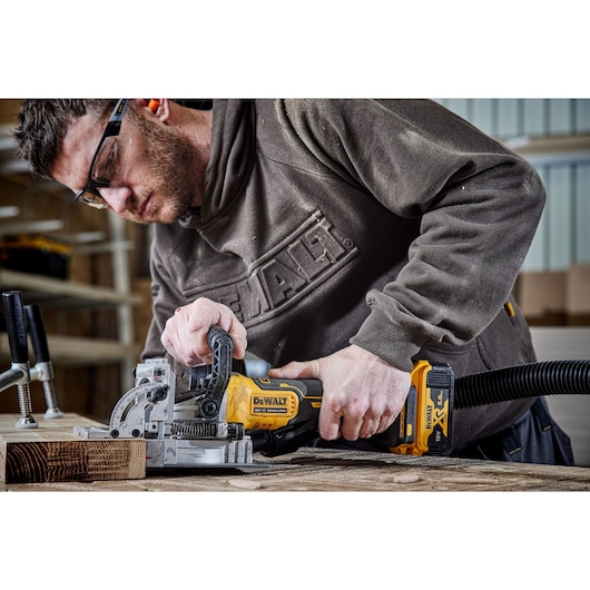 Person using a DEWALT DCW682N cordless jointer on wood in a workshop, attached to a dust extraction hose.