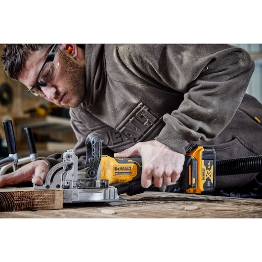 Person using a DEWALT DCW682N cordless biscuit joiner on a wooden workpiece, workshop setting.