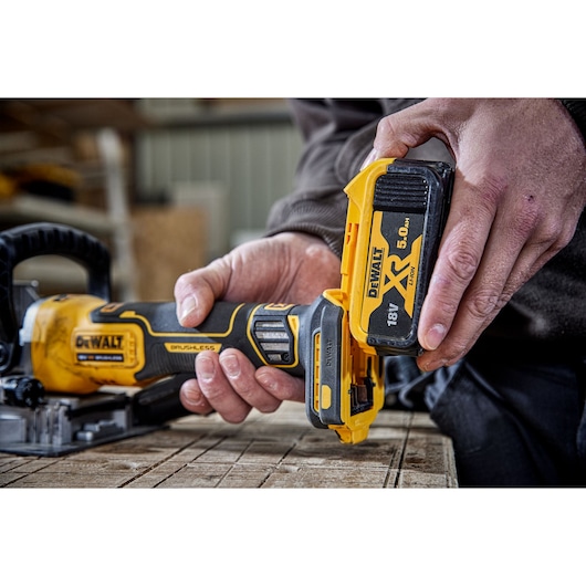 Close-up of DEWALT DCW682N tool with 18V XR battery being attached by a person at a woodworking workspace.
