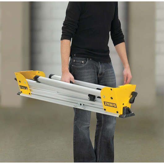 Person carrying a folded DEWALT DE7033 stand with yellow ends and metal frame indoors.