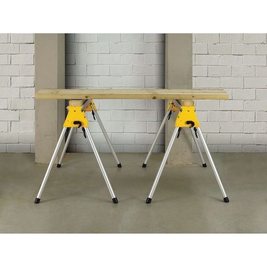 DEWALT DE7035 yellow metal work stand supports a wood plank, set against a concrete and brick wall indoors.