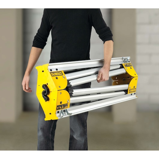 Person carrying a folded DEWALT DE7035 aluminum stand indoors.