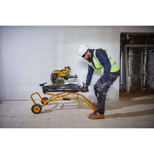 Worker moves a DEWALT miter saw on a yellow wheeled stand inside a construction site.