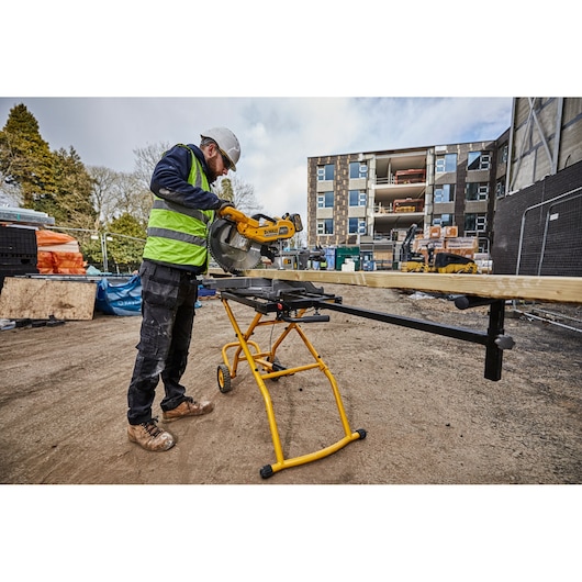 Worker uses DEWALT saw stand DE7260 to cut wood at a construction site. Building and equipment visible in background.